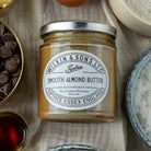 Jar of Wilkin & Sons Smooth Almond Butter surrounded by ingredients on a textured surface
