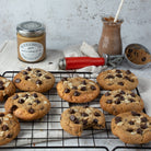 Chocolate chip cookies cooling on a wire rack, with a jar of Wilkin & Sons Almond Butter, a glass of chocolate sauce, and a red pastry scraper on a light stone surface.