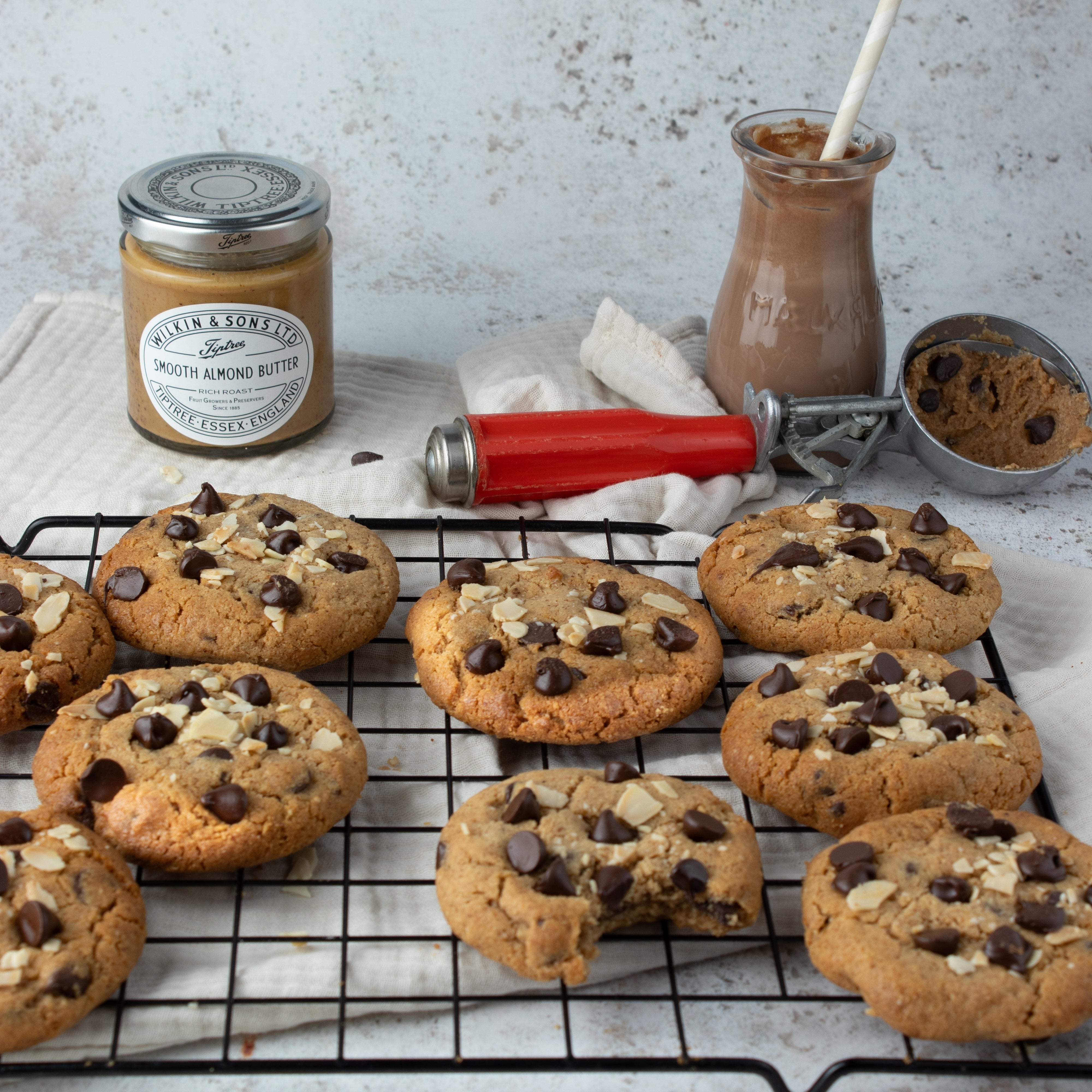 Chocolate chip cookies cooling on a wire rack, with a jar of Wilkin & Sons Almond Butter, a glass of chocolate sauce, and a red pastry scraper on a light stone surface.
