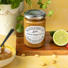 A jar of Wilkin & Sons Ltd. Crunchy Peanut Butter on a wooden board, styled with a bowl of peanut butter, whole peanuts, a lime half, and a potted cilantro plant against a warm yellow background.