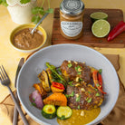 A plated dish of roasted chicken with vegetables in a creamy peanut sauce, served in a light blue bowl on a table with a jar of Wilkin & Sons Crunchy Peanut Butter, a small bowl of sauce, and garnish.