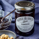 A jar of Wilkin & Sons Ltd. Damson Conserve, styled on a dark blue textured cloth with a glass bowl of jam and whole hazelnuts.