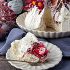 A slice of meringue cake with damson jam and figs on a plate in the foreground, with the full cake in a fluted dish behind it. A jar of damson jam and plums are visible on a dark rustic background.