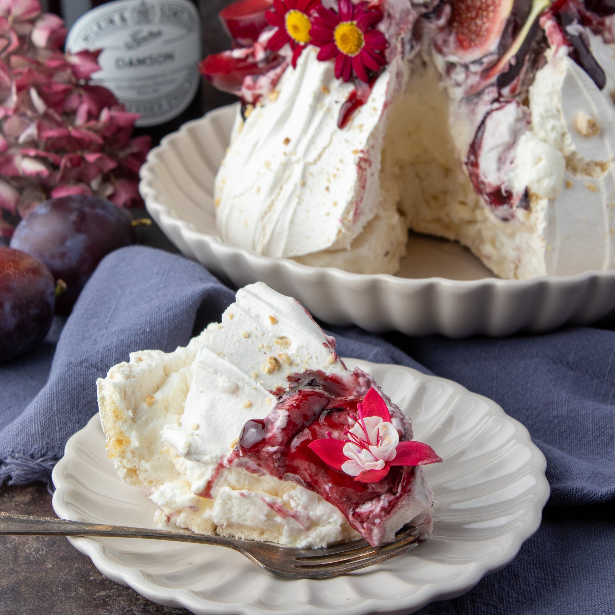 A slice of meringue cake with damson jam and figs on a plate in the foreground, with the full cake in a fluted dish behind it. A jar of damson jam and plums are visible on a dark rustic background.