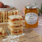 A jar of Wilkin & Sons Ltd. Tiptree Peach Conserve alongside stacks of peach jam shortbread crumble bars on a styled tabletop with peaches in the background.