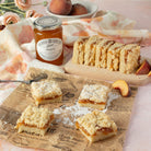 Peach jam shortbread crumble bars  arranged on brown printed newspaper with a jar of peach jam and whole peaches in a styled food scene.