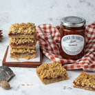A jar of Wilkin & Sons Quince Conserve surrounded by baked oatmeal bars with jam filling, styled on a white textured surface with a red-and-white checkered cloth, a grater, and a nut.