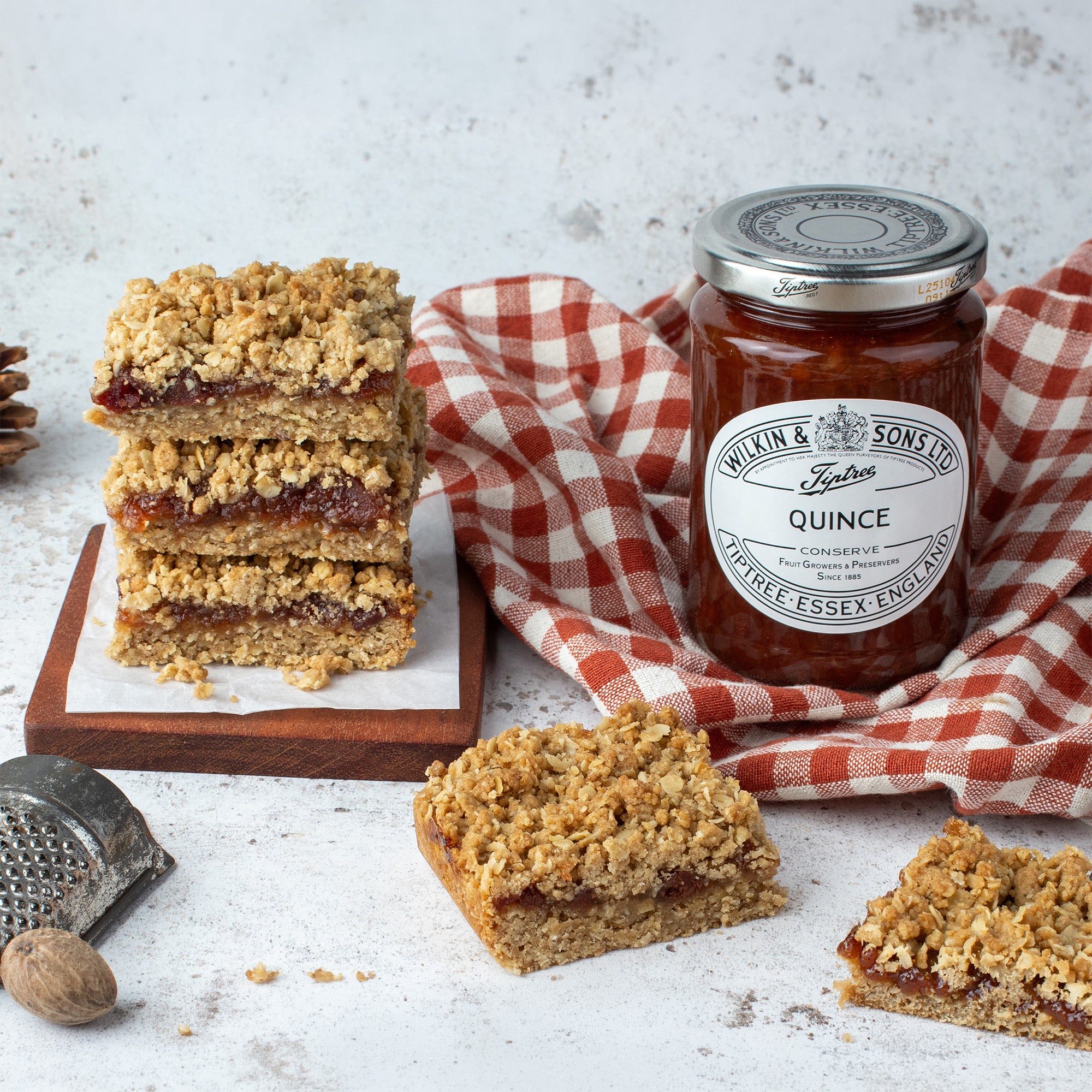 A jar of Wilkin & Sons Quince Conserve surrounded by baked oatmeal bars with jam filling, styled on a white textured surface with a red-and-white checkered cloth, a grater, and a nut.