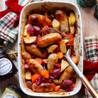 Baked sausages with vegetables in a white dish on a wooden table.