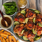 Overhead food scene showing glazed chicken pieces on a plate with lime wedges and red chili slices, served with bowls of marinade, broccoli, potato wedges, and a jar of sauce on a rustic wooden table.