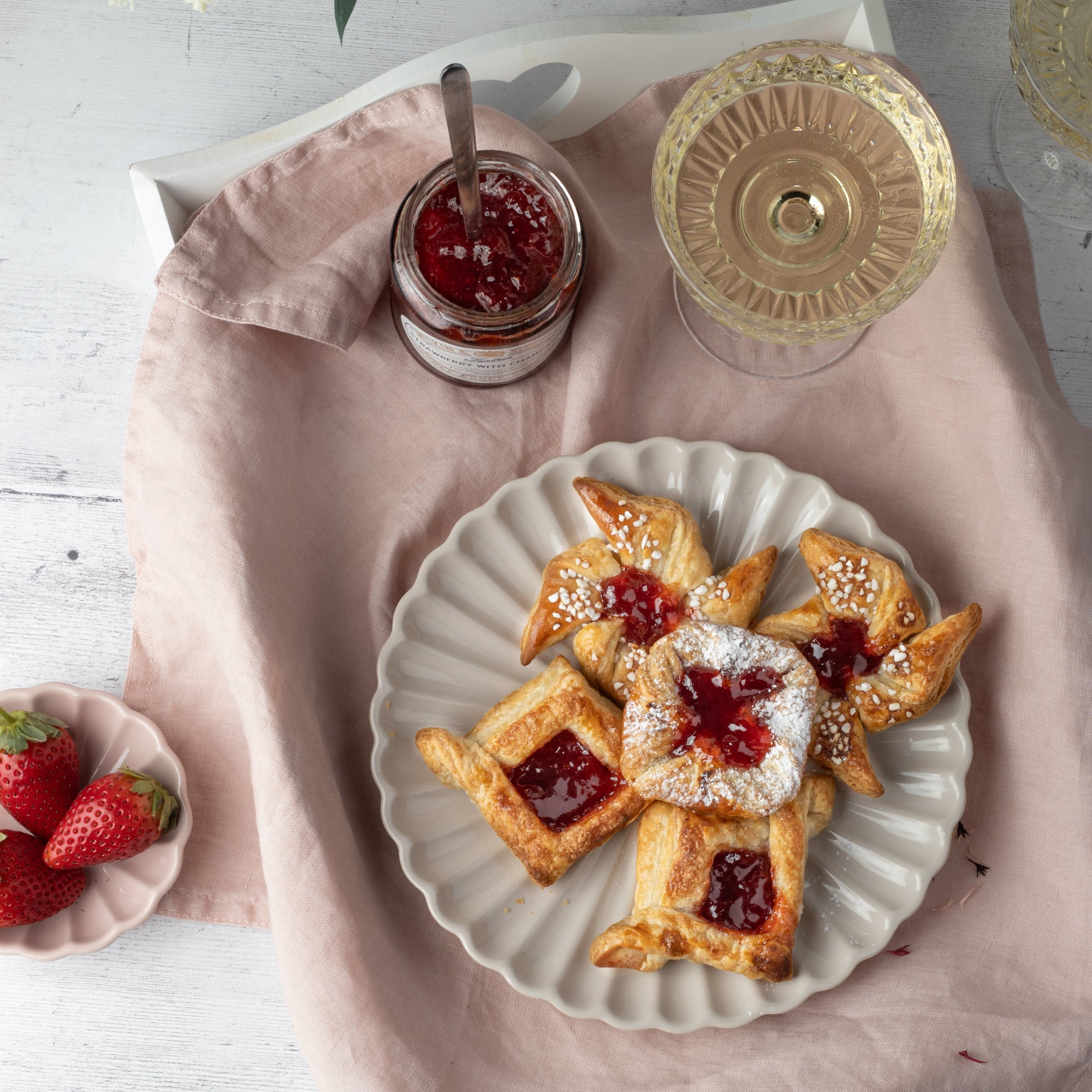 A styled food scene showing several golden flaky pastries topped with strawberry jam, dusted with powdered sugar, and arranged on a white plate over a pink cloth. A jar of jam with a spoon sits nearby, along with strawberries and a glass on a white tabletop.
