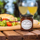 Jar of Tiptree Chutney on a wooden table with a blurred background of food and glasses.